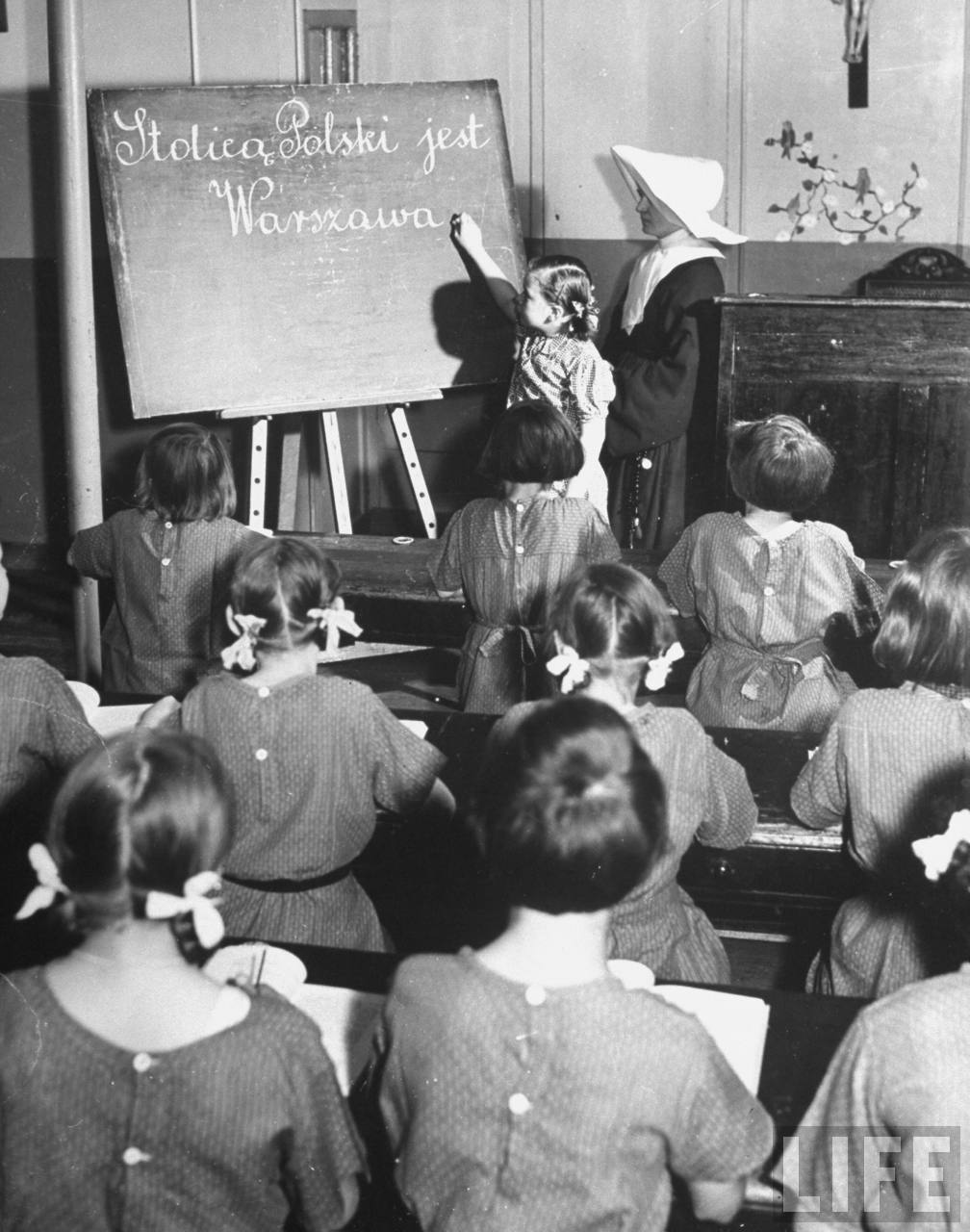 A girl working at the chalkboard of a classroom while a nun and her classmates look on.