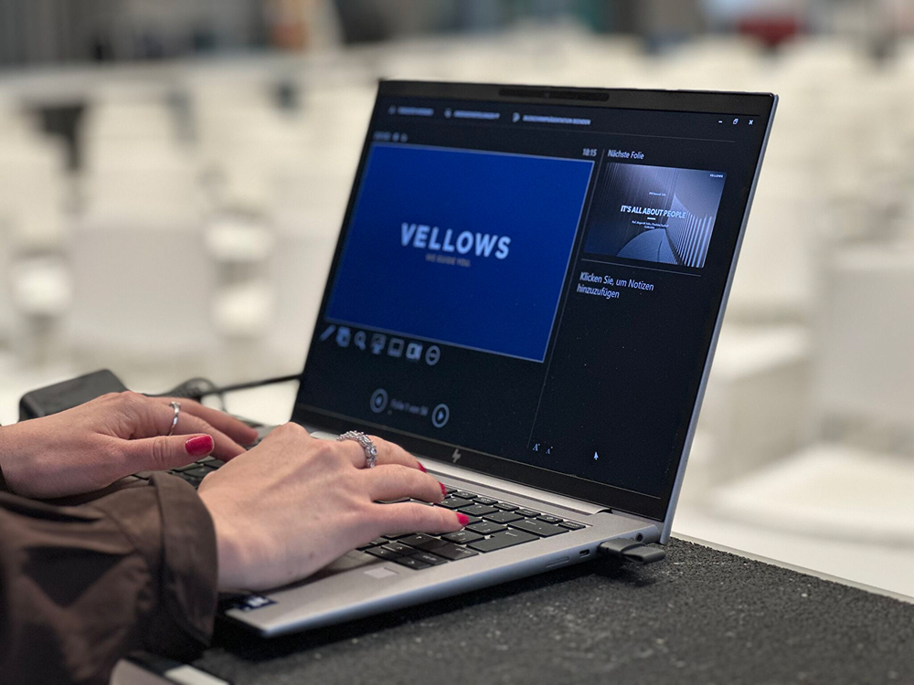 Close-up of a person with rings typing on a laptop displaying a "VELLOWS" presentation in a blurred, bright room