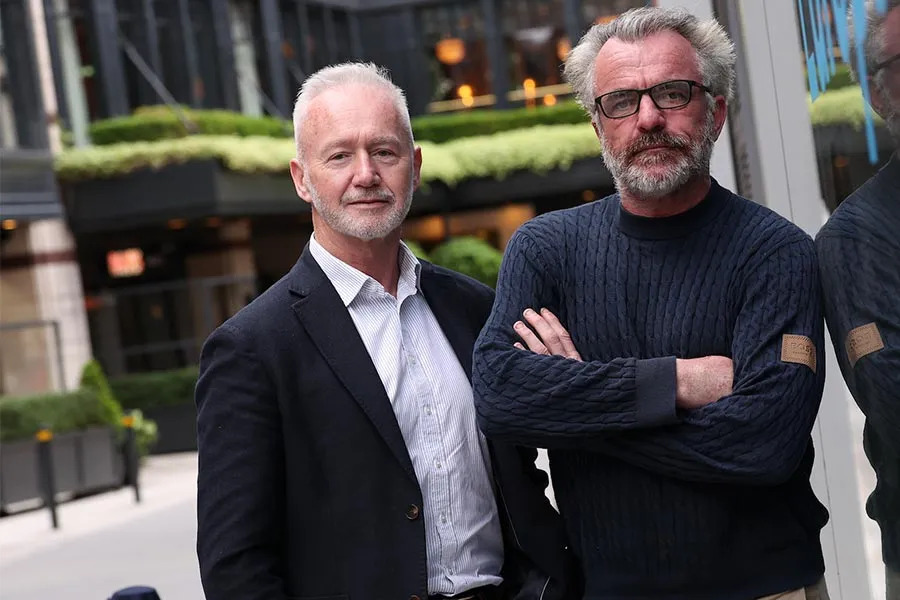 Two men, one in a blazer and one in a cable-knit sweater, posing outdoors in front of a modern building