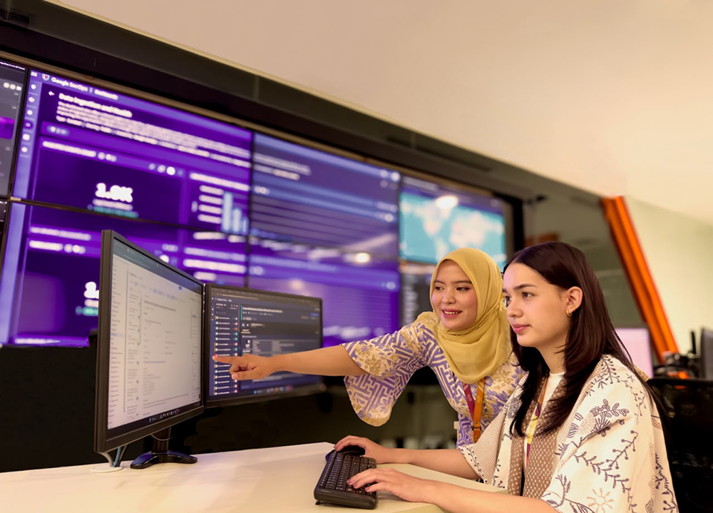 Two individuals working in an office environment with computer monitors and a large display screen in the background