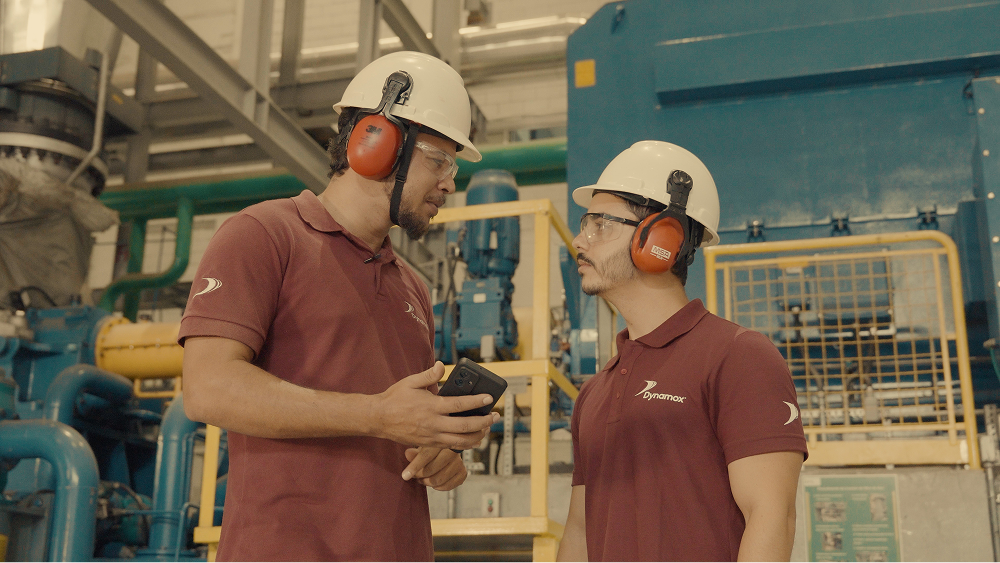 Dois trabalhadores com capacetes e protetores auriculares conversam em uma fábrica, um deles segurando um celular