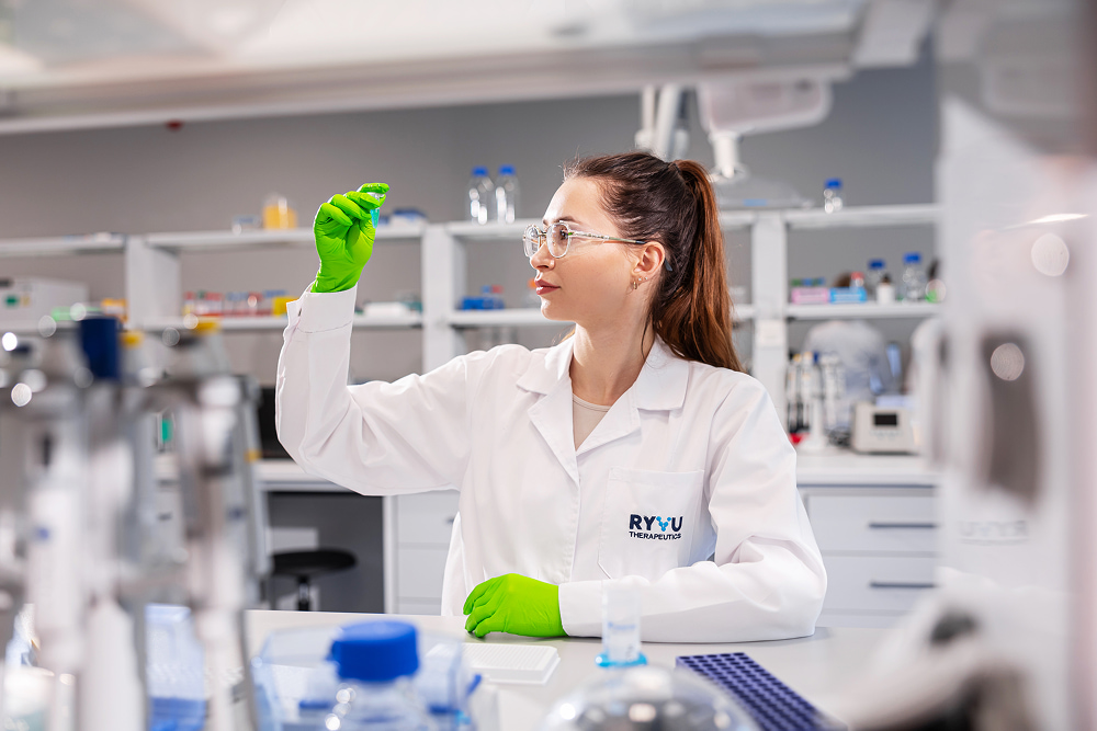 Scientist in a lab coat and green gloves examines a sample at Ryvu Therapeutics, surrounded by medical equipment