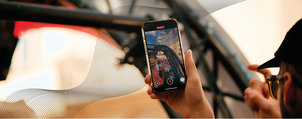 A person's hands holding a smartphone to record a video of a DJ performing at a crowded outdoor event through a metal frame