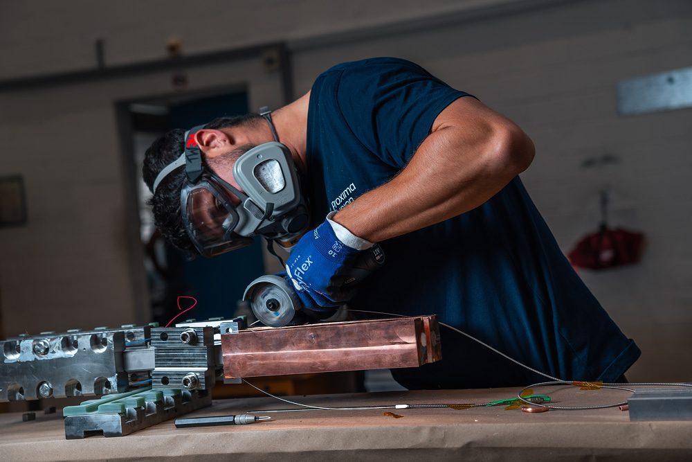 A man wearing a respirator, safety goggles, and blue gloves uses an angle grinder on a copper workpiece in a workshop