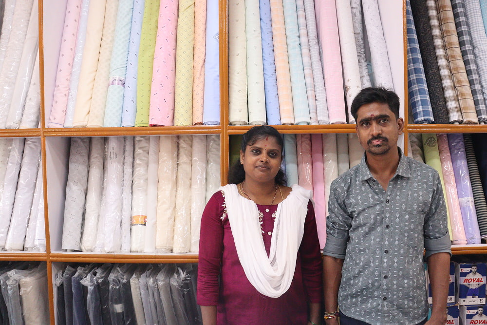A man and woman stand in front of shelves filled with colorful fabric bolts in a shop. Some boxes labeled "ROYAL" are visible