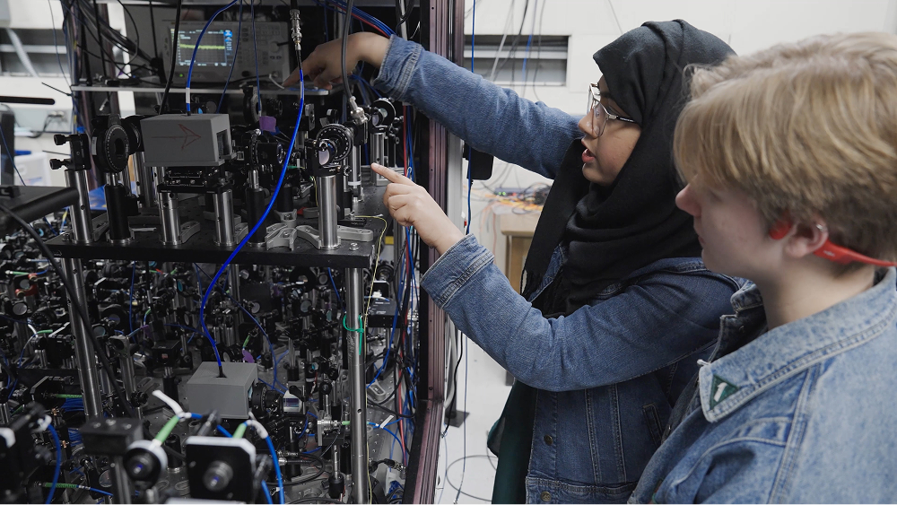 Two students work in an optics laboratory, with one wearing a hijab pointing at a complex setup of equipment