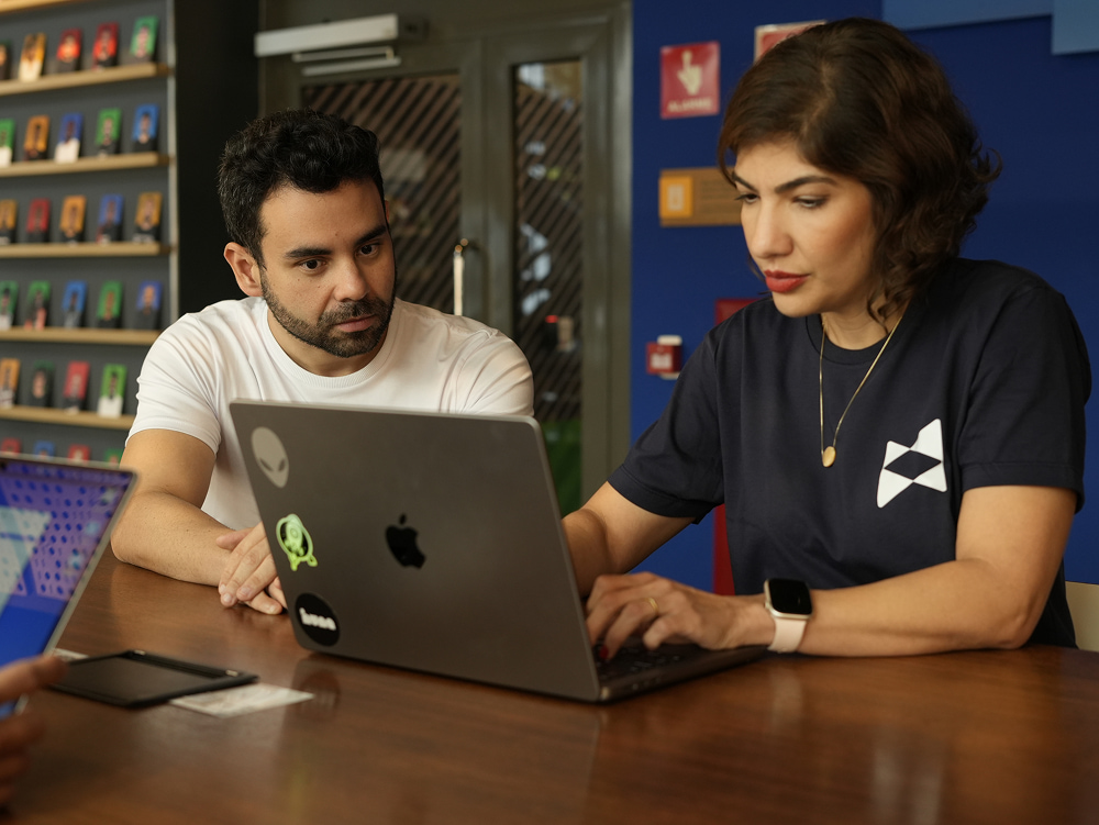 Vinicius e Daniella, fundadores da Huna, olham para a tela de um laptop enquanto trabalham juntos em uma mesa de madeira