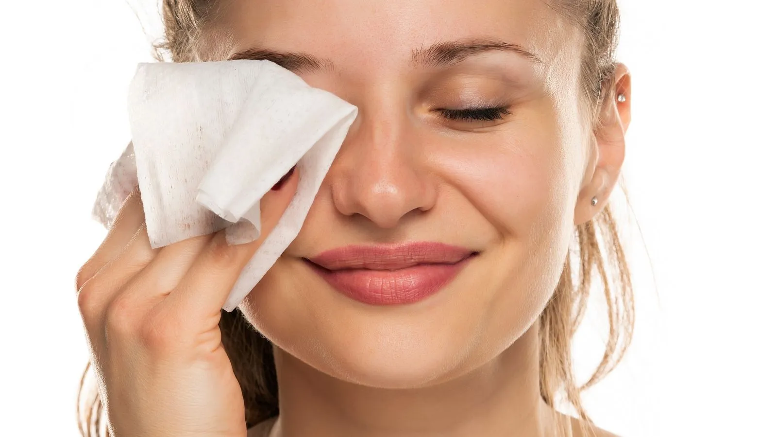 A woman smiling with her eyes closed while using a white makeup remover wipe on her face against a white background