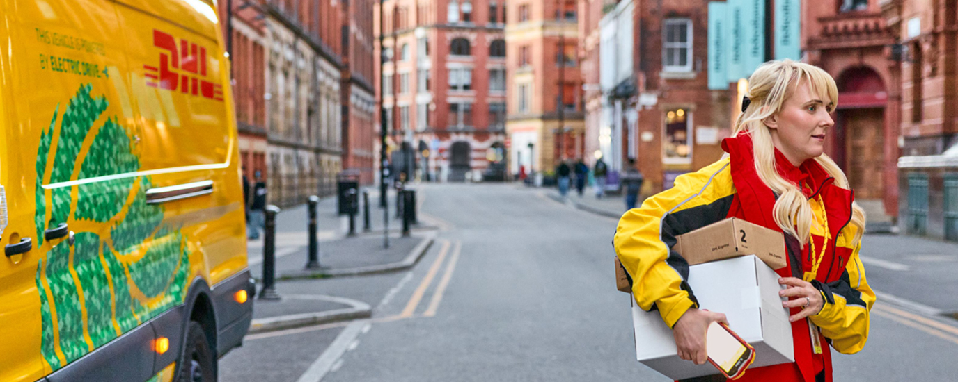 A DHL delivery driver carrying packages walks down a city street next to a yellow electric DHL delivery van