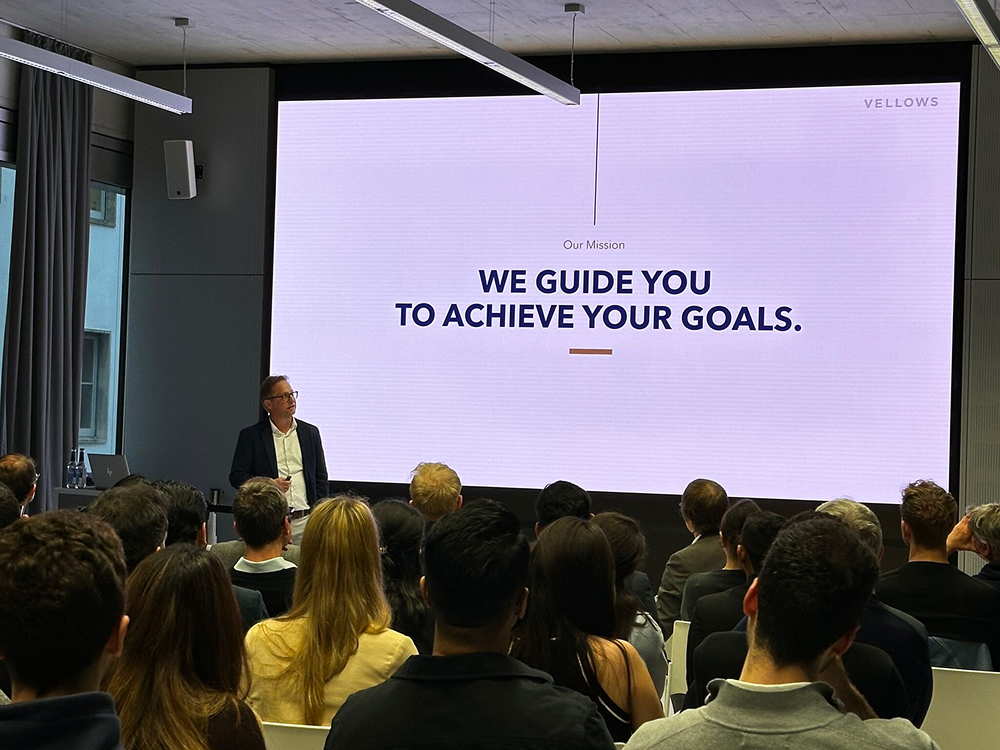 A man gives a presentation to an audience; the screen reads "Our Mission WE GUIDE YOU TO ACHIEVE YOUR GOALS"