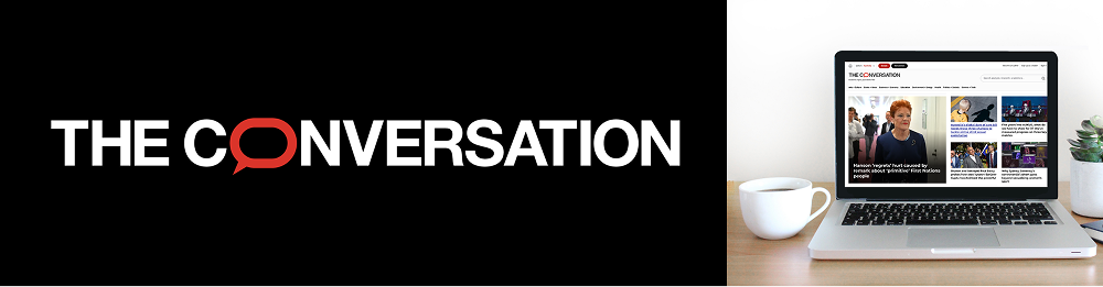 A laptop displaying "THE CONVERSATION" website next to a white coffee mug on a desk, with the site's logo on the left