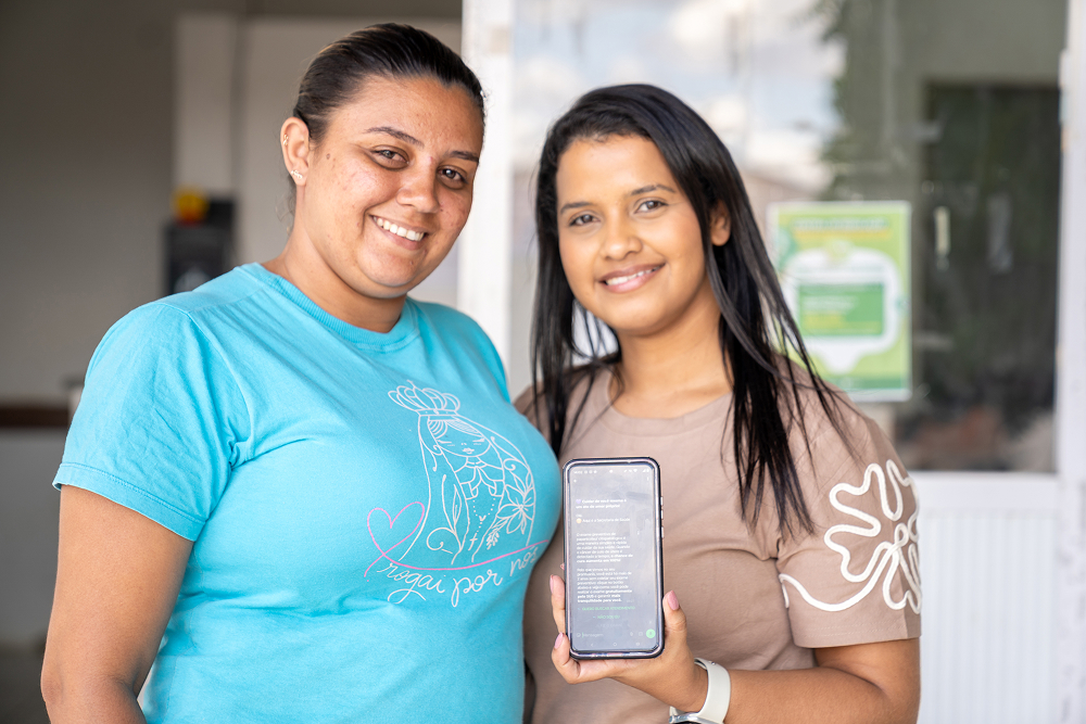 Duas mulheres sorriem lado a lado; a da direita segura um celular mostrando uma tela de texto
