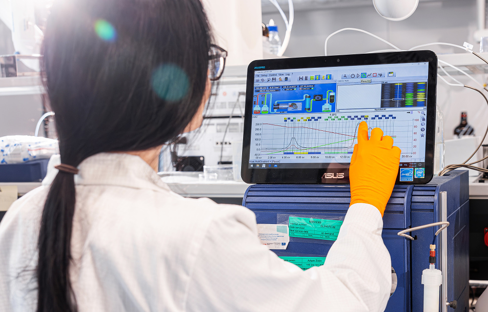 A scientist in a white lab coat and orange gloves touches a digital screen displaying complex data graphs in a laboratory