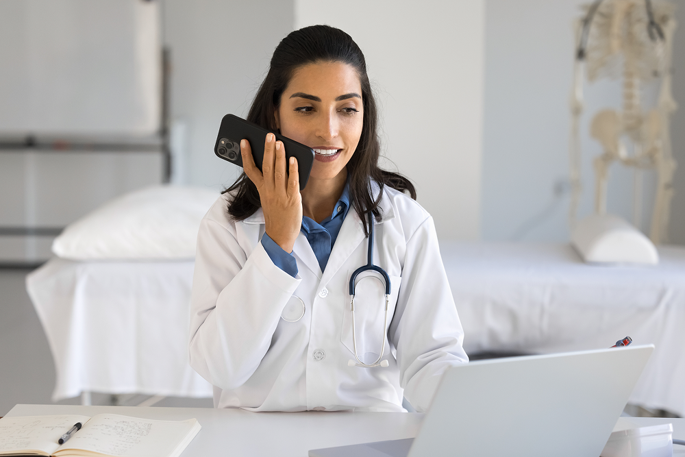 A female doctor wearing a white lab coat and stethoscope speaks into her smartphone while working at a laptop in her office
