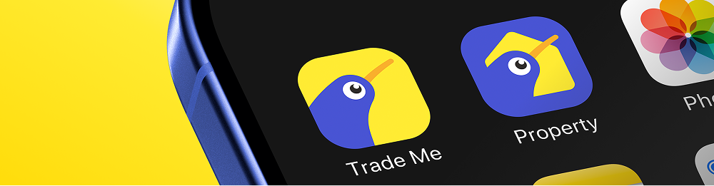 Close-up of a blue smartphone screen showing the Trade Me and Property app icons, featuring a stylized kiwi bird logo