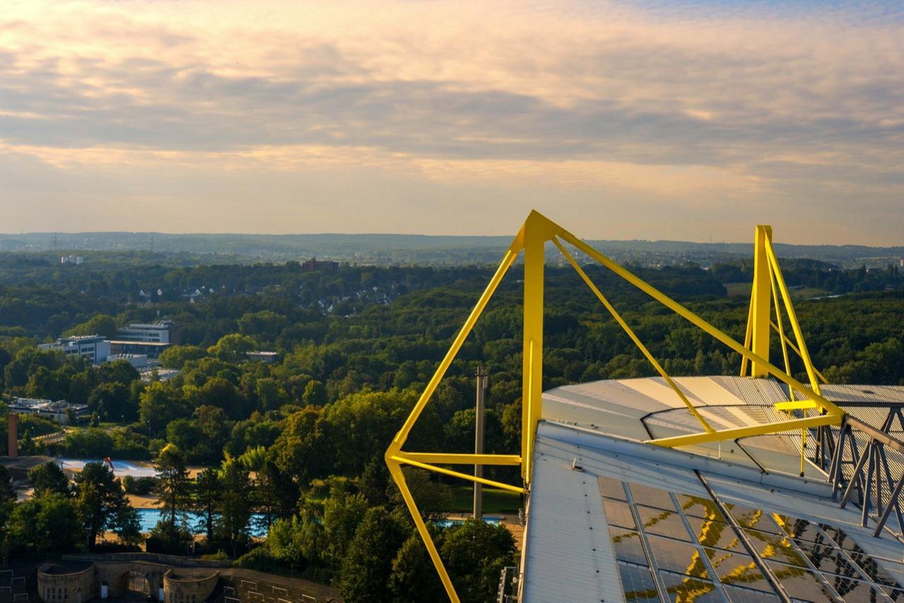 High-angle view of yellow steel stadium pylons overlooking a sprawling green forest landscape under an evening sky