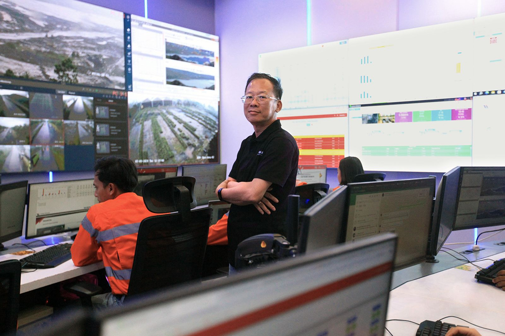 Man in glasses stands in control room surrounded by screens