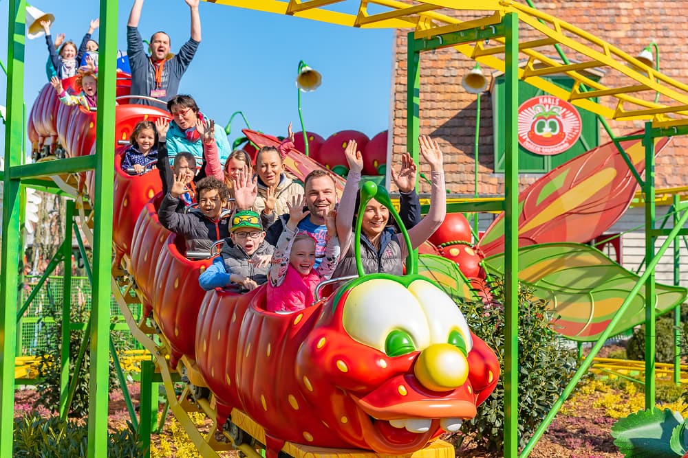 A vibrant scene at an amusement park or fair, showing a group of people riding a caterpillar-themed roller coaster