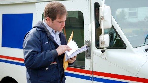 postal employee reviews pieces of mail in front of USPS vehicle