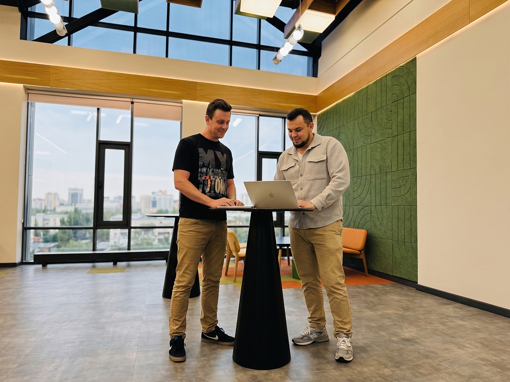 Two men stand at a high-top table in a modern office, collaborating over a laptop in front of large windows