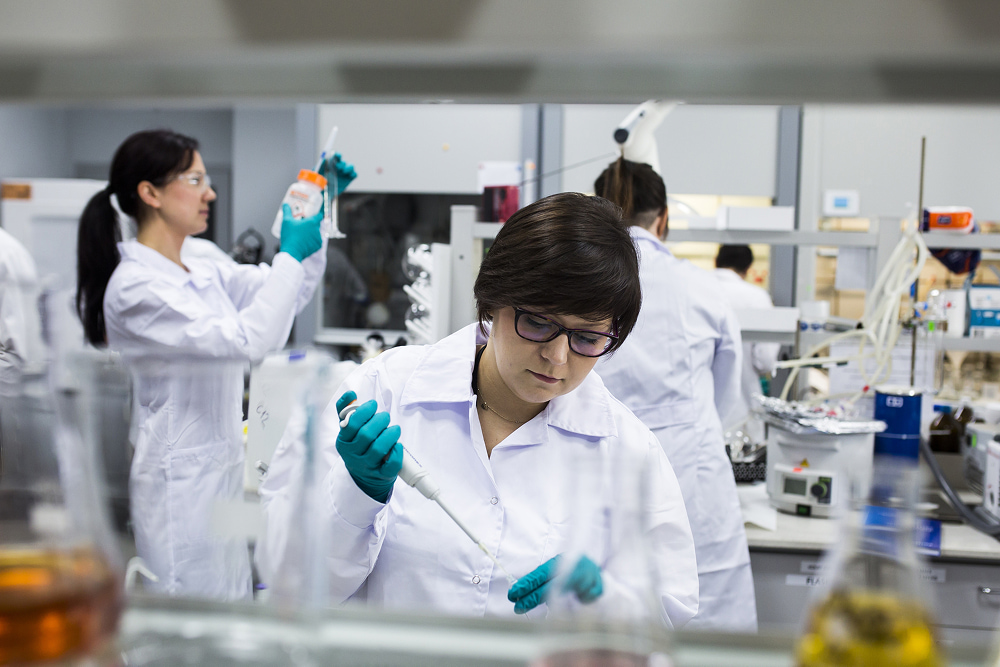A female scientist in a white lab coat and blue gloves uses a pipette in a busy laboratory with colleagues