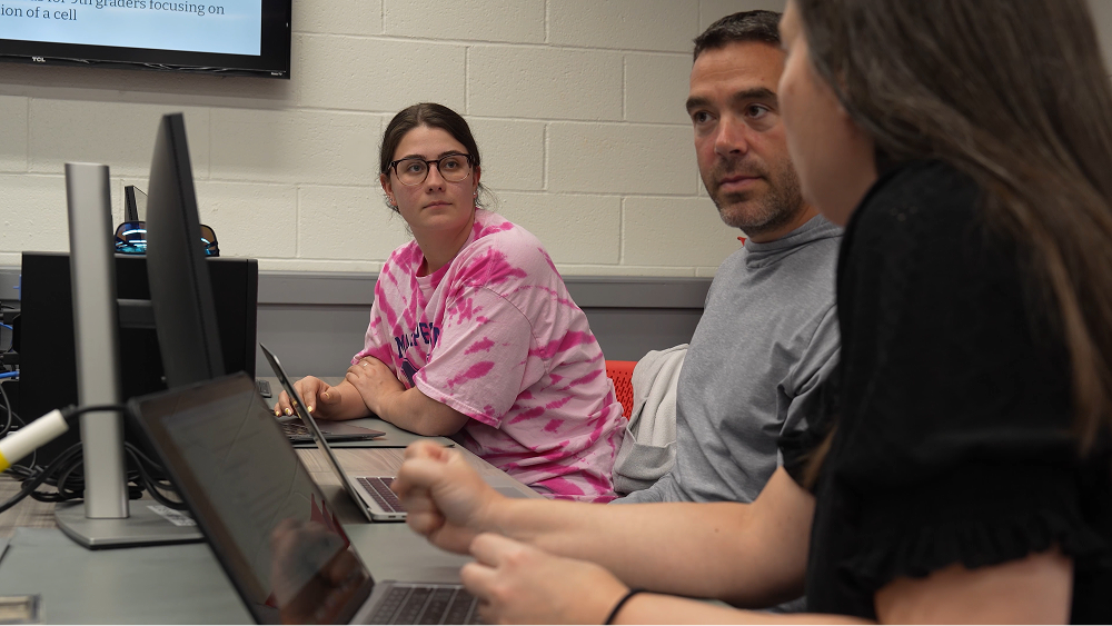 A woman in a pink tie-dye shirt and a man in a grey shirt look on as a colleague works on a laptop in a classroom