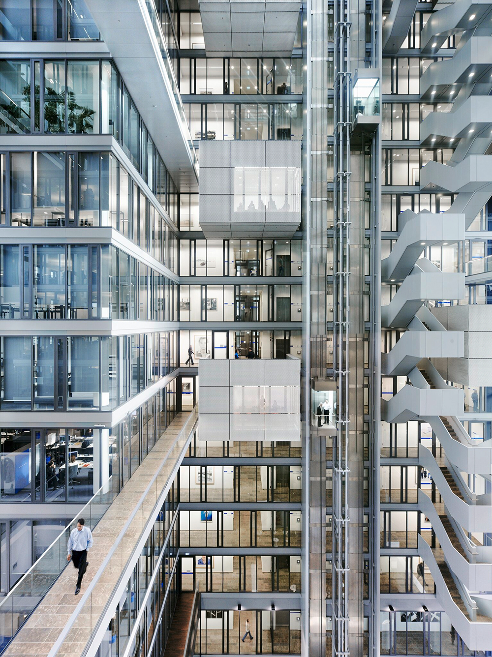 A multi-story modern office atrium with glass walls, suspended walkways, elevators, and a zigzagging staircase