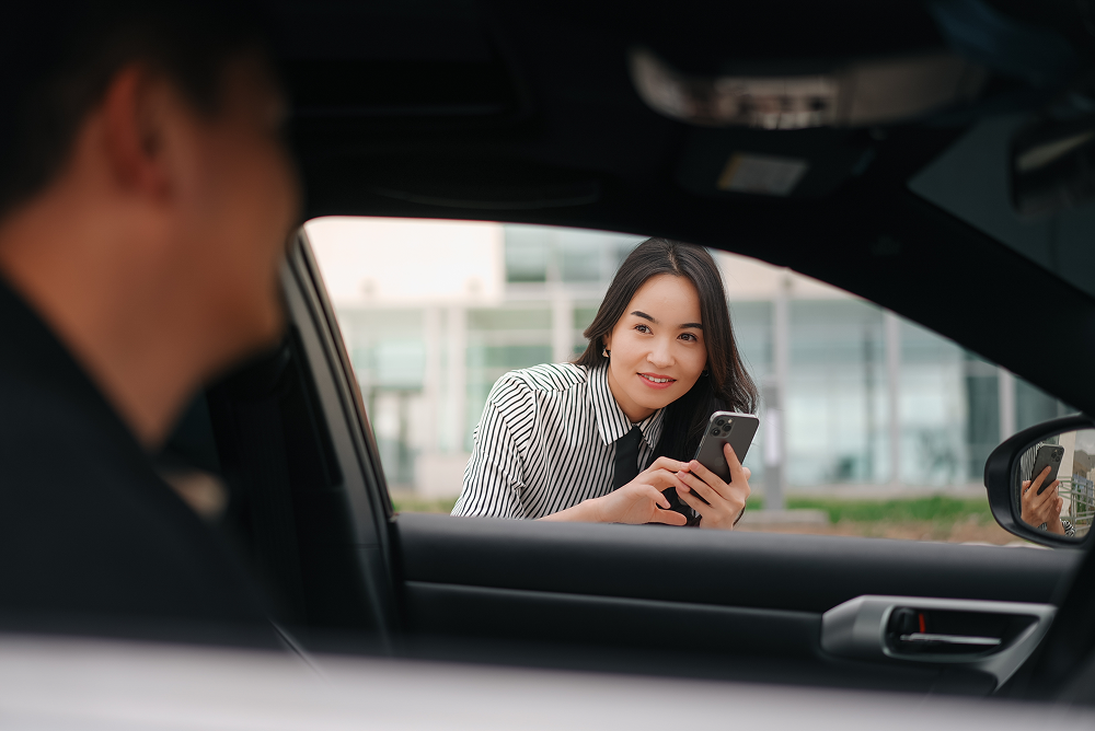 A person looking out from inside a vehicle at a woman standing outside while holding a smartphone