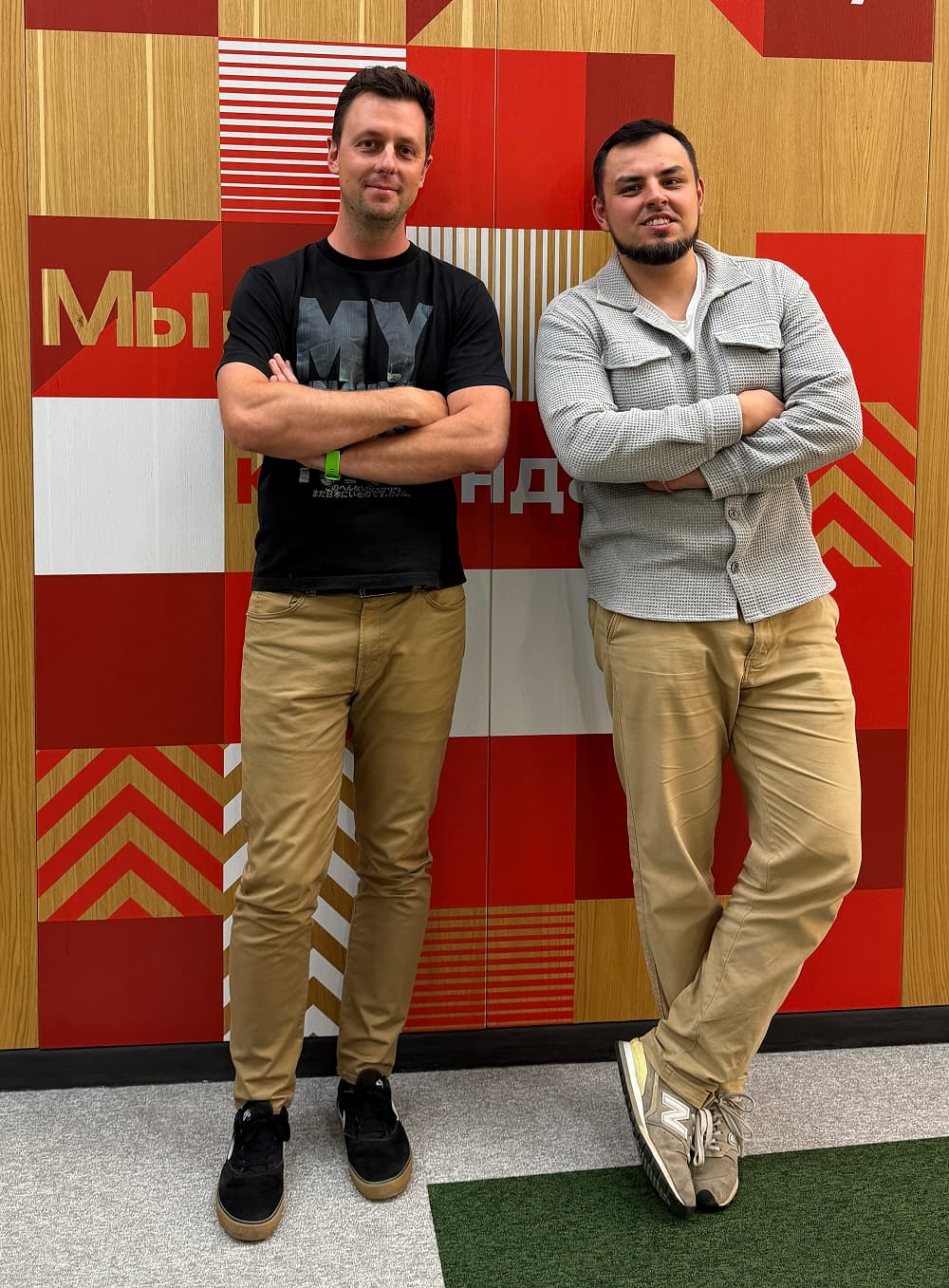 Two men stand with arms crossed against a red and gold geometric wall