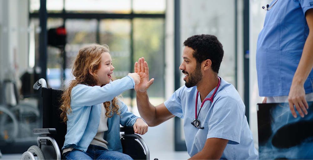 A smiling male nurse in blue scrubs high-fives a young girl sitting in a wheelchair in a brightly lit hospital hallway