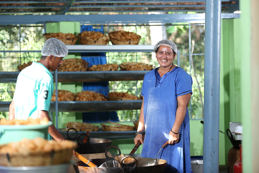 Woman in a blue dress and man in a light green shirt, both wearing hairnets, cook food in a large kitchen