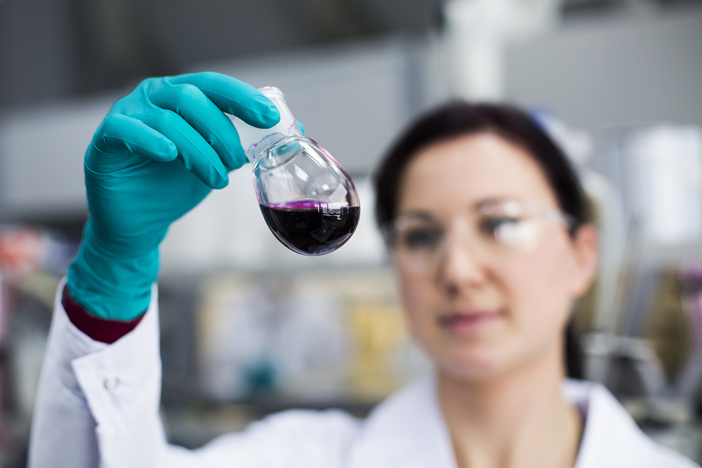 Focused shot of a scientist in a lab coat and green gloves holding up a glass flask filled with dark purple liquid