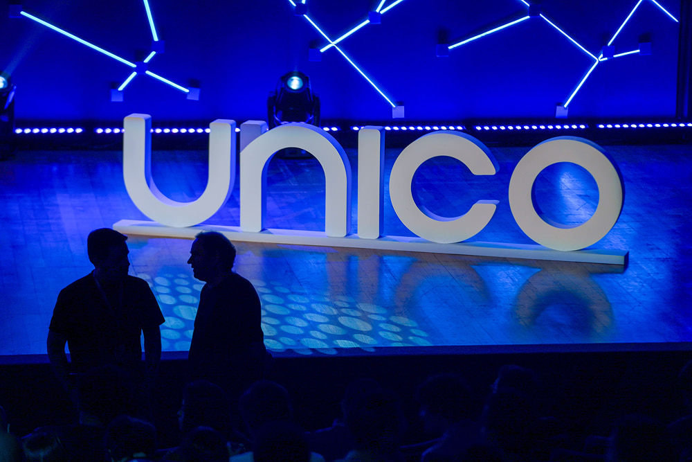 Two silhouetted figures standing on a stage in front of a large, illuminated sign that reads "unico" with neon lights