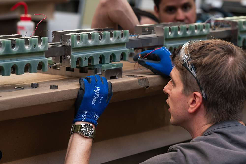 Worker in safety glasses and blue gloves carefully assembles complex industrial machinery on a manufacturing line