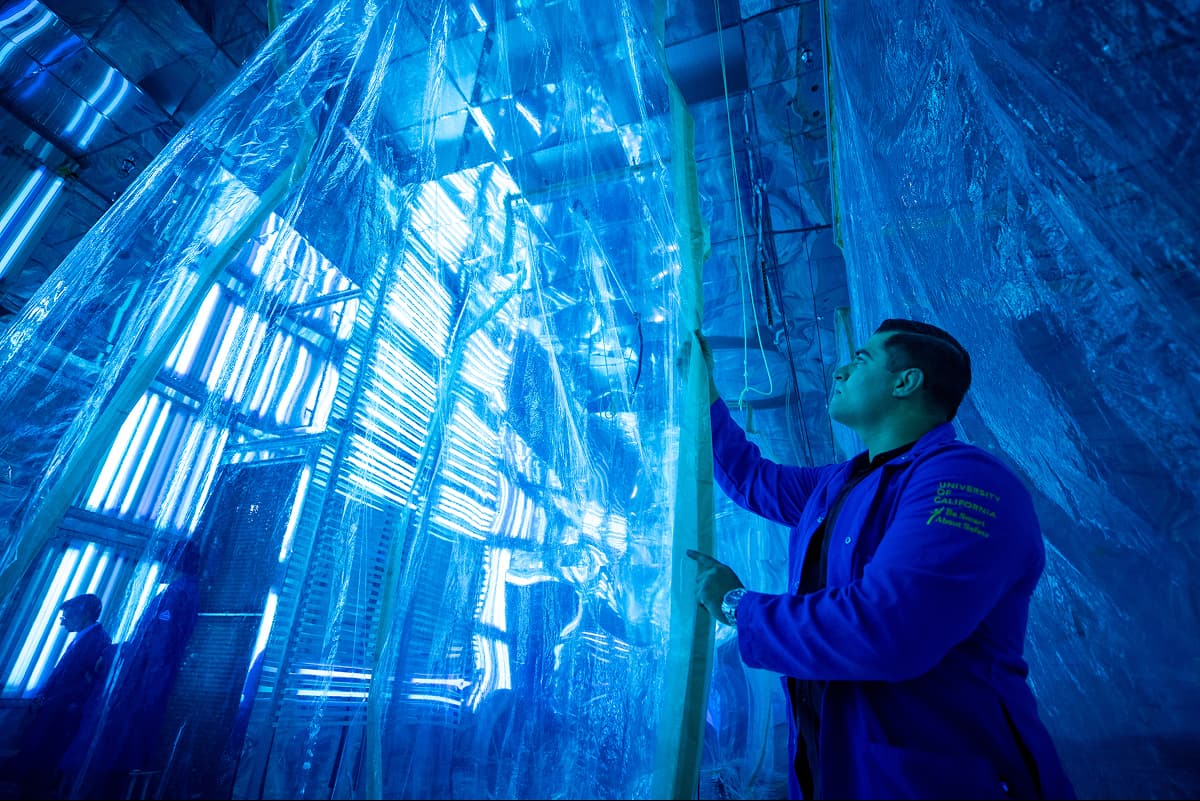 Man in a bright blue lab coat reaching up to adjust or examine a large, transparent blue plastic sheet or structure.
