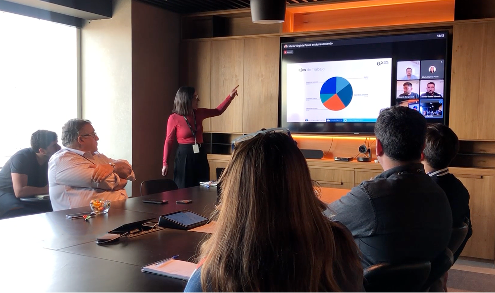 A woman stands in a conference room pointing at a pie chart displayed on a large screen during a hybrid business meeting