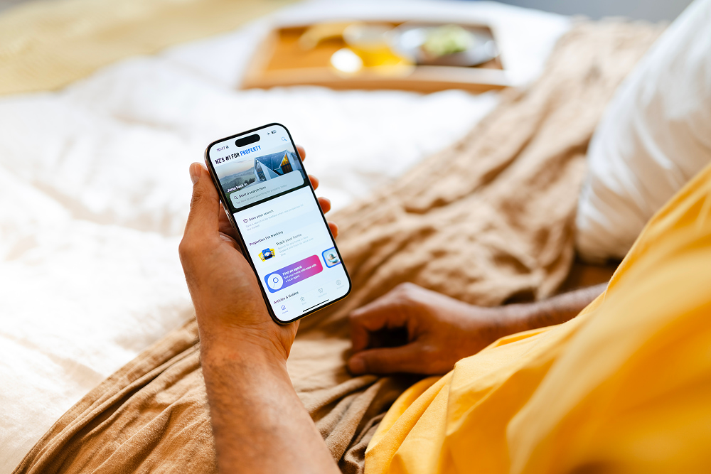 A person in a bed holds a smartphone showing a real estate app, with a breakfast tray blurred in the background.