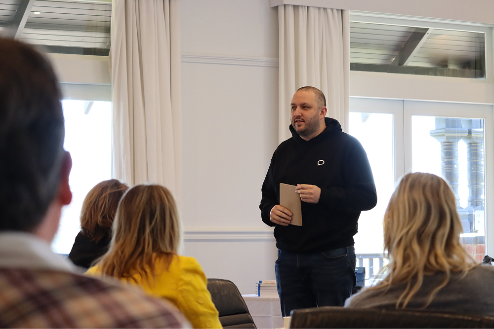 A speaker in a black hoodie holds a card, standing in front of an audience inside a brightly lit room during a presentation