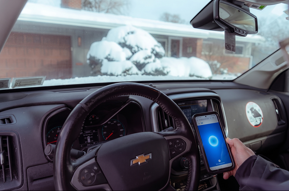 A person in a Chevrolet holds a phone displaying Gemini Live, while a snow-covered house is visible through the windshield