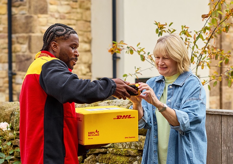 A DHL Express delivery person hands a handheld device to a smiling woman for a signature while holding a yellow package