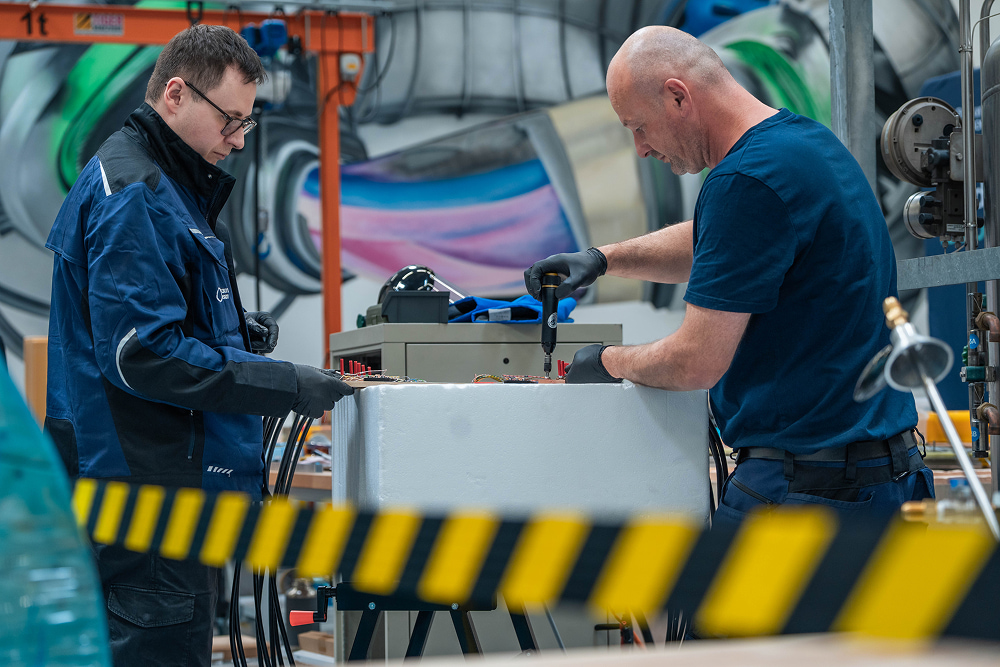 Two men in a workshop assemble components on a white block