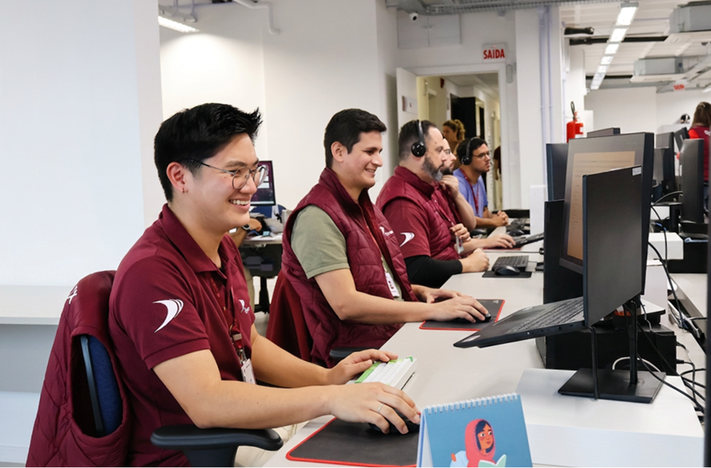 Pessoas trabalhando em um escritório com computadores, vestindo coletes e camisas polo na cor vinho e sorrindo