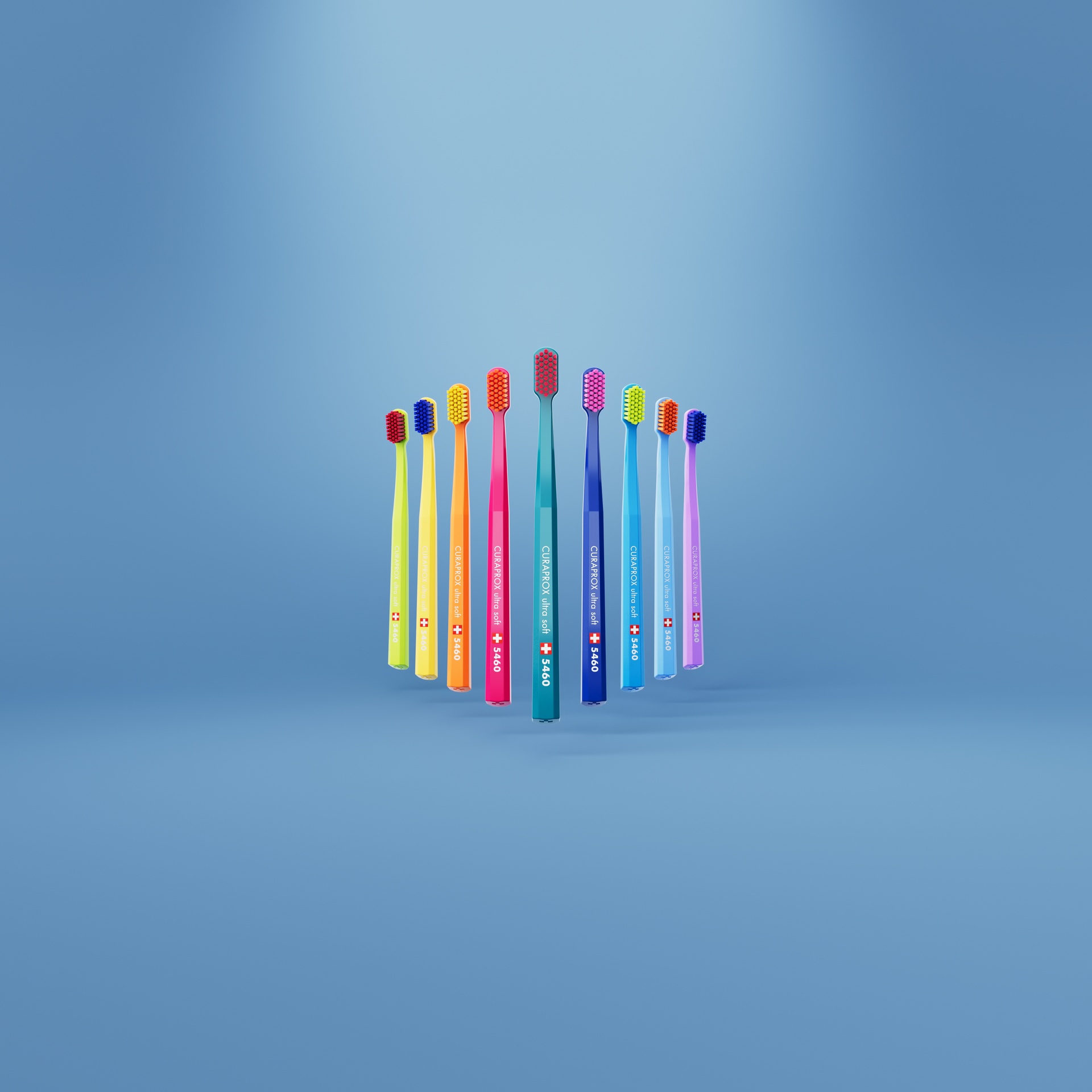 Twelve colorful Curaprox ultra soft 5460 toothbrushes arranged in a floating circle against a soft blue background