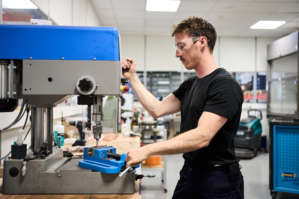 A man wearing safety glasses operates a large blue and gray industrial drill press on a workbench in a bright workshop