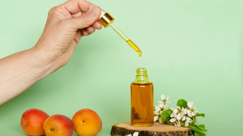 Hand holding a dropper with oil over a glass bottle, with apricots and white flowers on a light green background