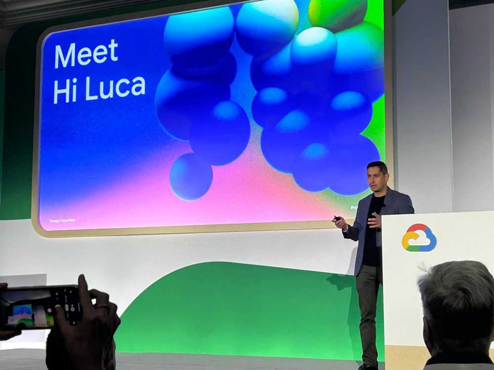 A presenter on stage at Google Cloud Next standing next to a large screen that displays "Meet Hi Luca" on a blue background