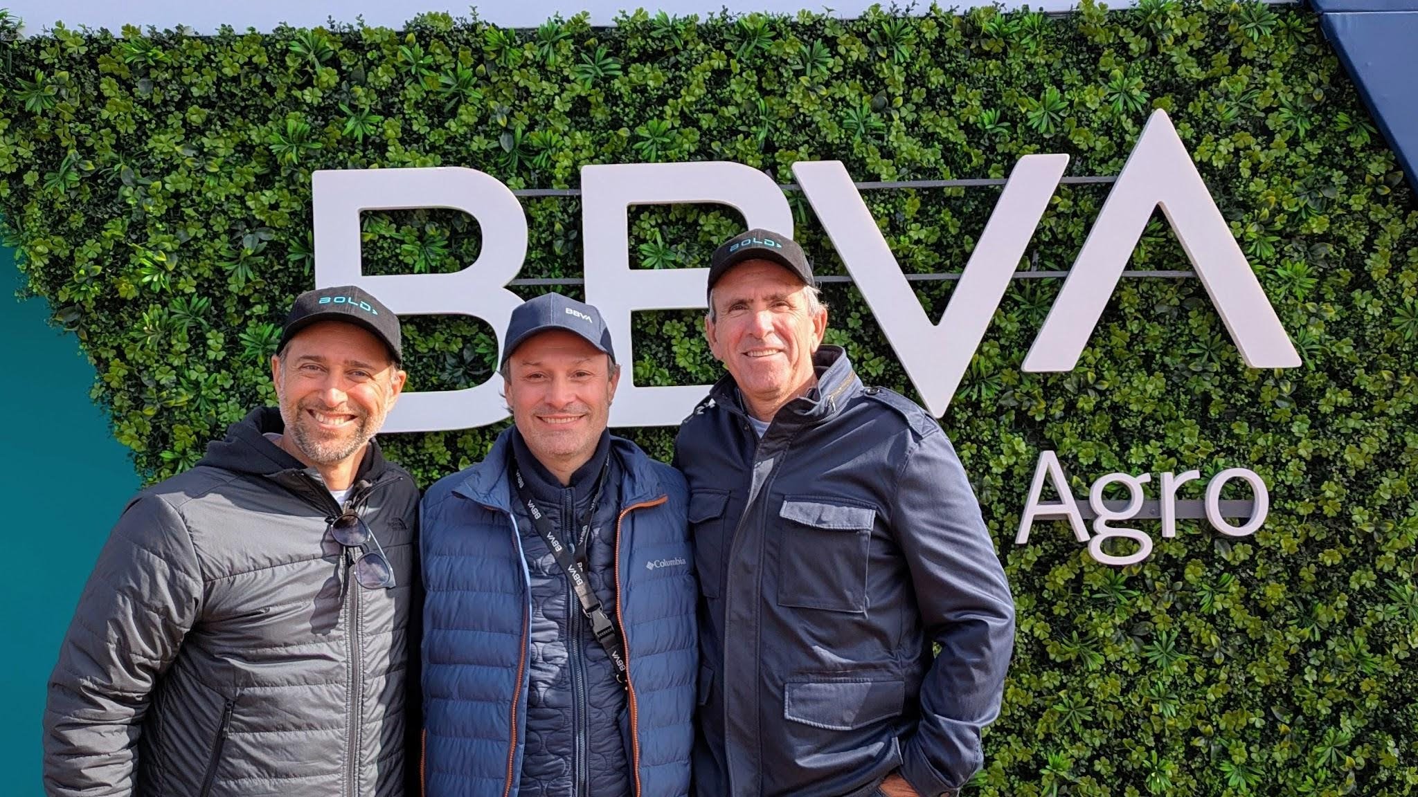 Three men smile in front of a green hedge wall featuring the large white letters "BBVA Agro" 