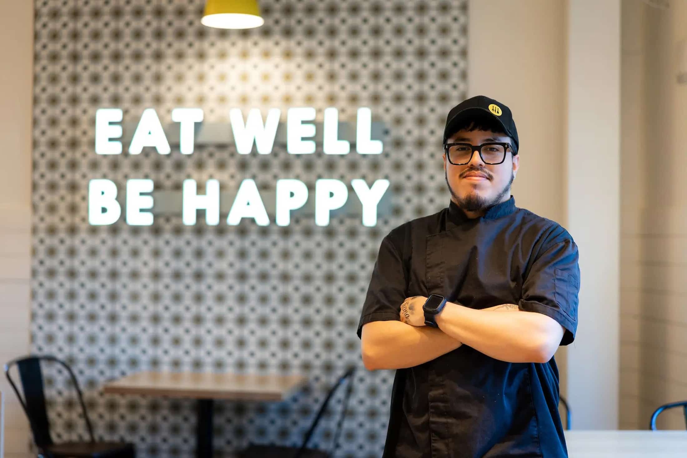 Chef in black uniform posing by "Eat Well Be Happy" sign
