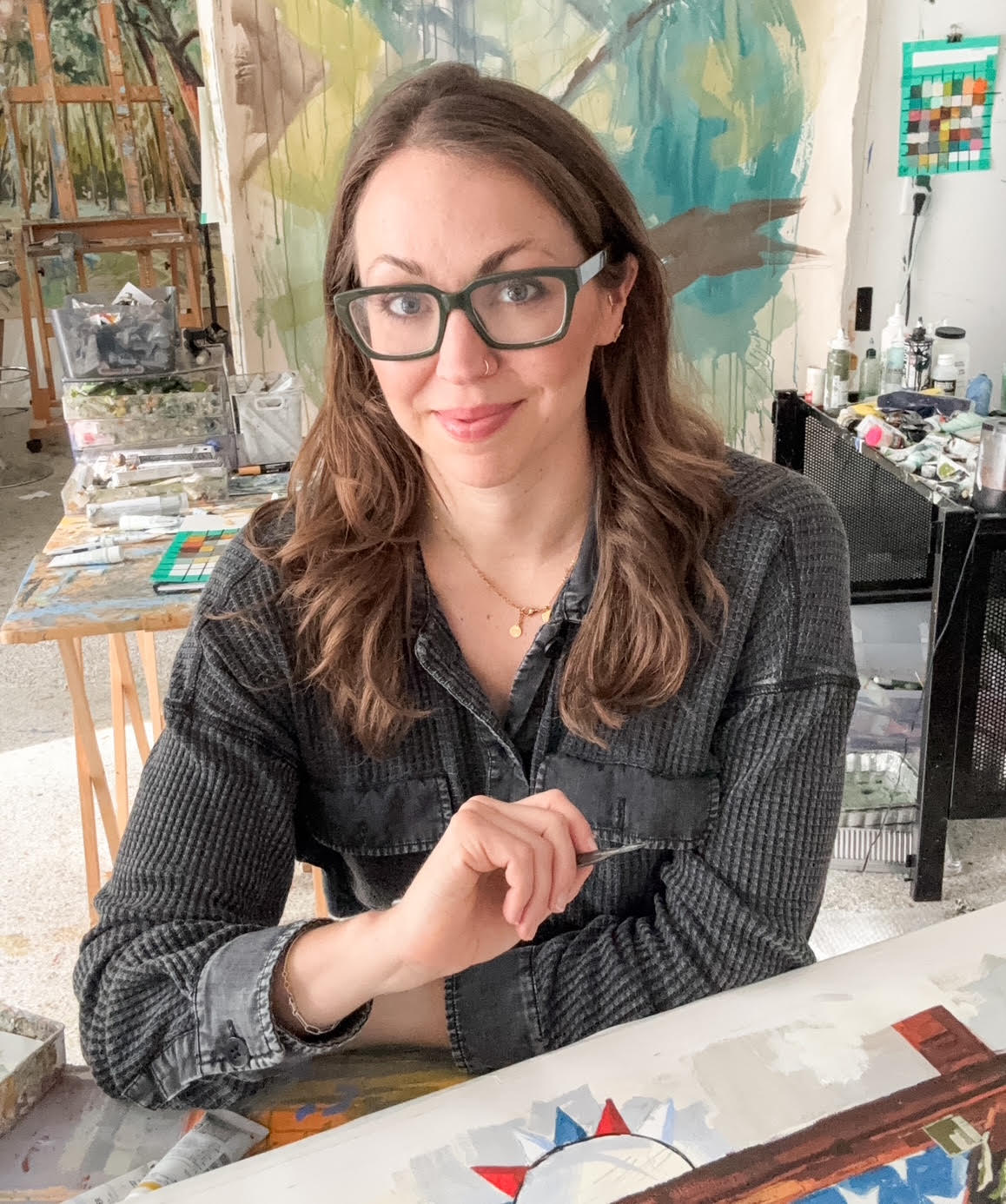 Photograph of a white woman with dark hair and eyeglasses sitting in an art studio