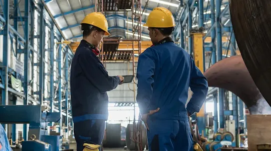 Two industrial workers in yellow hard hats and blue uniforms collaborating on a digital tablet inside a large manufacturing facility.